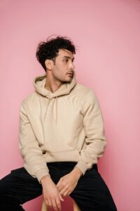 Thoughtful male with dark hair in stylish clothes sitting on wooden chair and looking away in studio against pink background