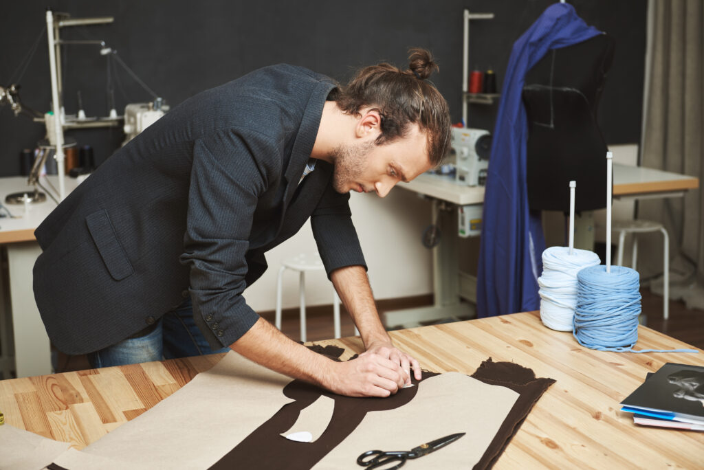 portrait of manly good looking adult male clothes designer with stylish hairstyle in black suit cutting out parts of future dress from fabric. man concentrated on work.